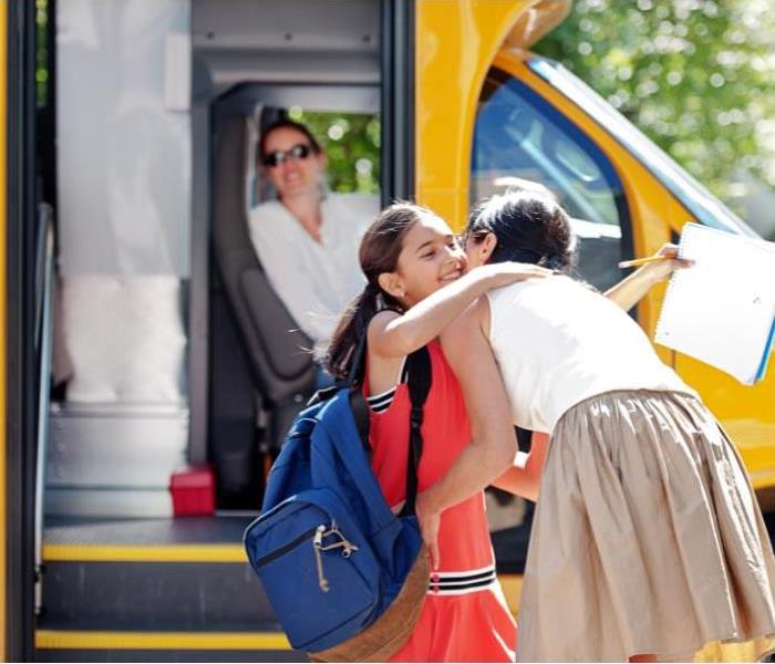 School kids going back to school. Little girl hugging her mom with a school bus in the background. 